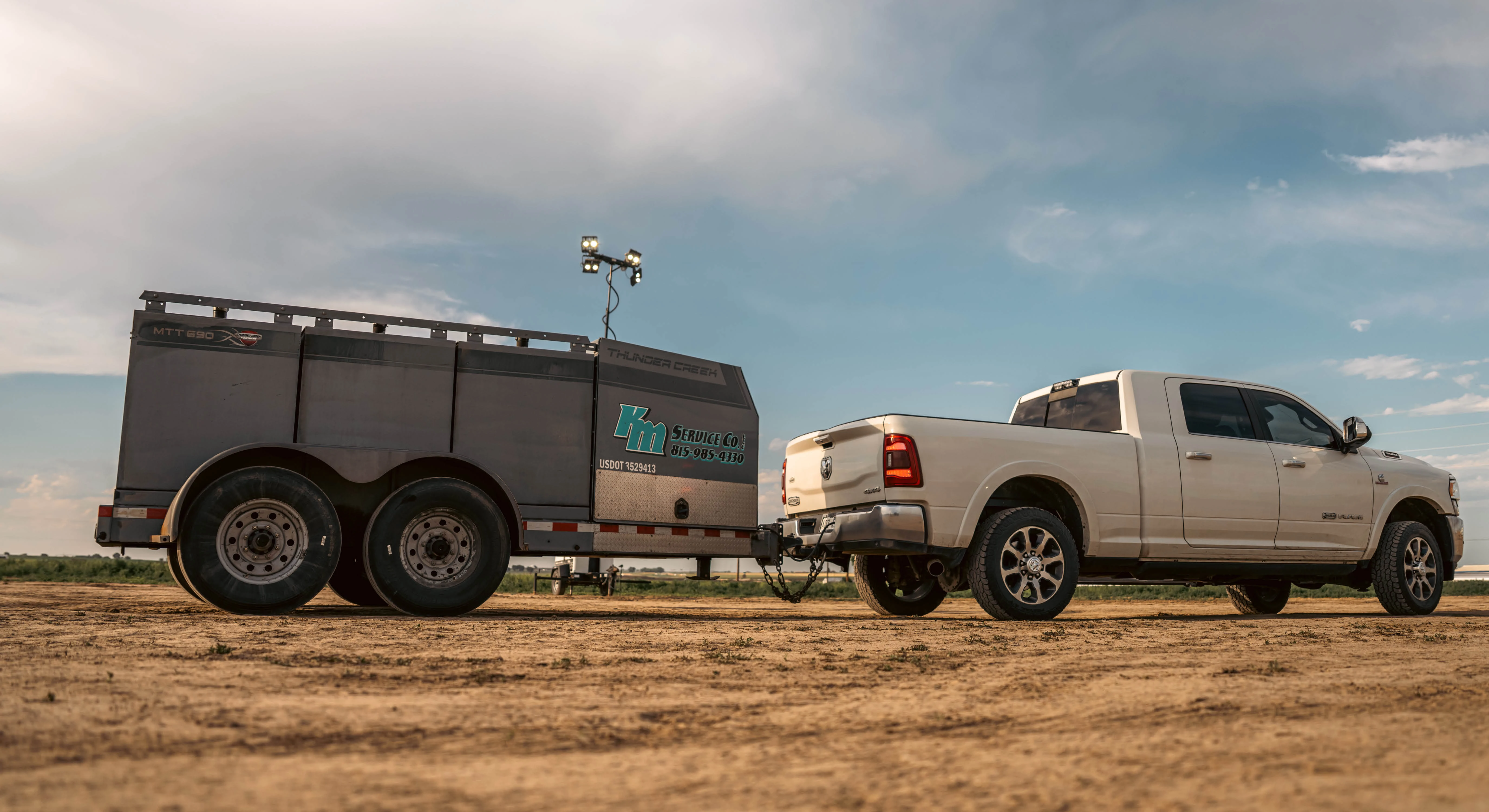 A white pickup truck tows a Thunder Creek Equipment Multi Tank Trailer with KM Service Co. decals on a dirt field under cloudy, blue skies.